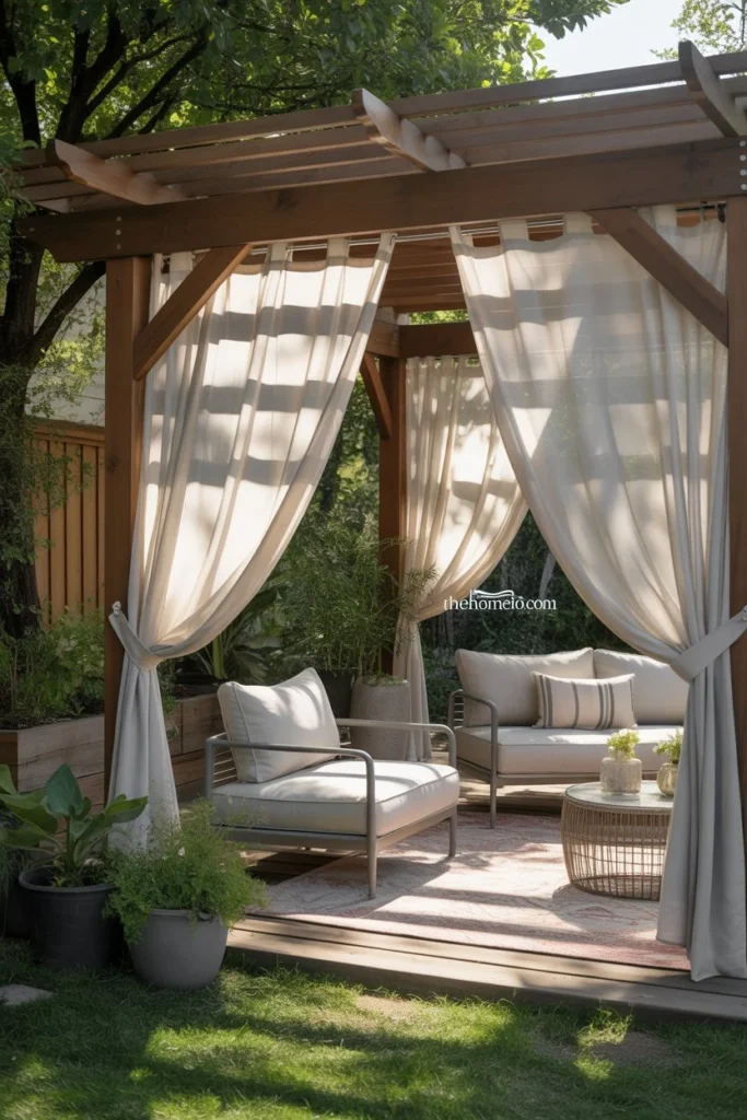 A modern pergola patio with sheer white curtains gently billowing in the breeze, neutral-colored lounge chairs, soft beige outdoor rug, greenery surrounding the patio, sunlight filtering softly, cozy and airy atmosphere, realistic American backyard